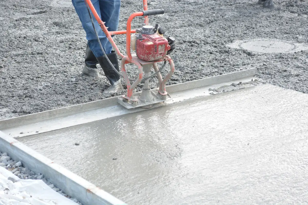 Worker running a vibratory power screed along steel forms across a freshly placed concrete slab