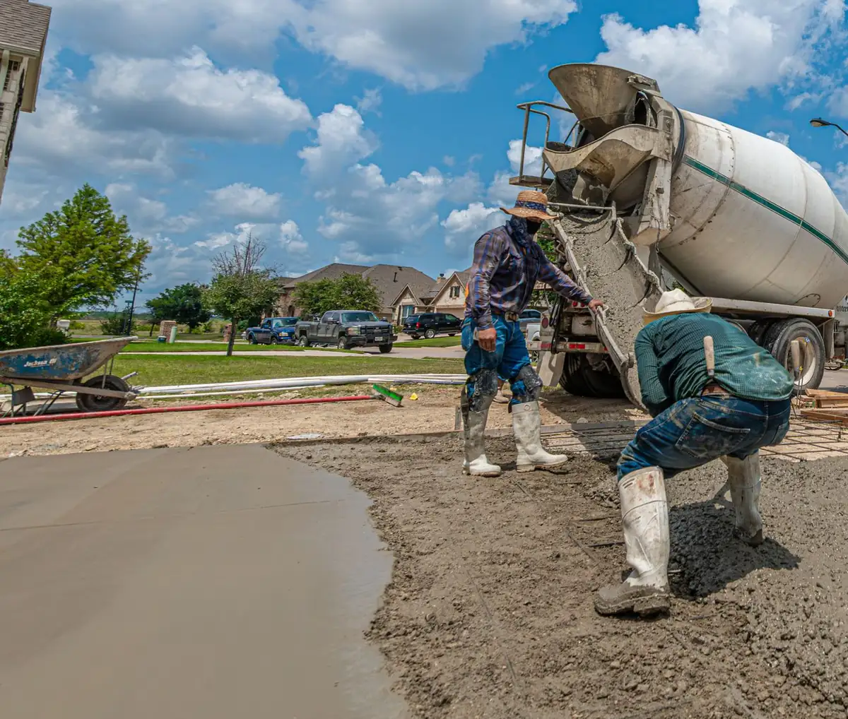 Ready-mix truck chute placing concrete over wire mesh on a residential driveway base while a finisher spreads it
