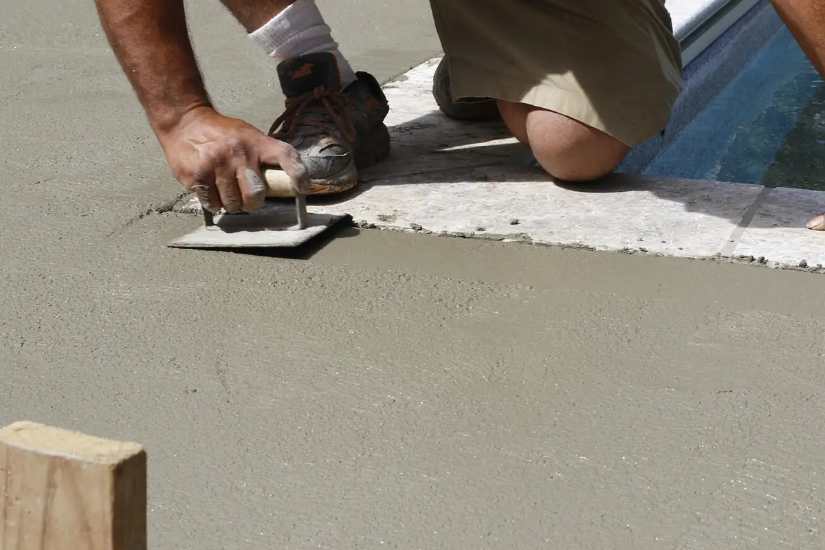Finisher kneeling beside a pool coping, smoothing a concrete deck surface with a hand float